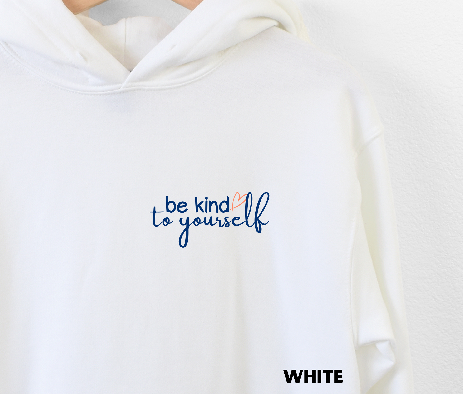 Close up of an embroidered Hoodie in white with "be kind to yourself' in navy blue text with a pink heart on the upper left chest. 