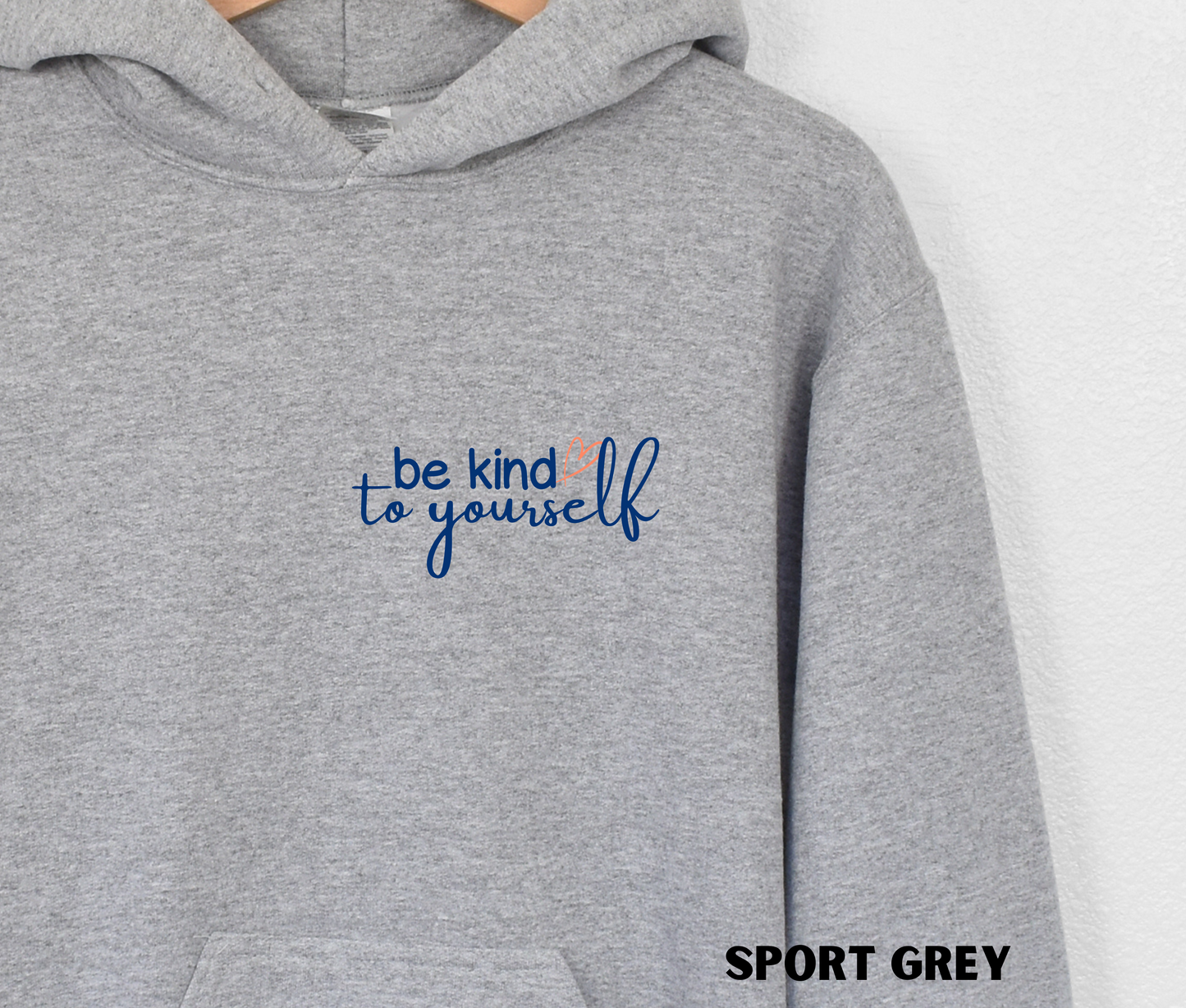 Close up of an embroidered Hoodie in sport grey with "be kind to yourself' in navy blue text with a pink heart on the upper left chest. 