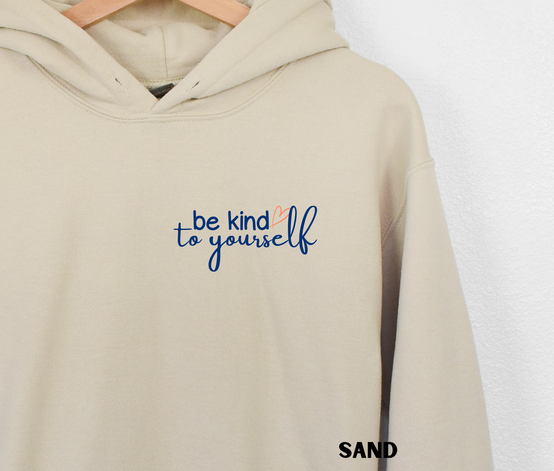 Close up of an embroidered Hoodie in a sand color with "be kind to yourself' in navy blue text with a pink heart on the upper left chest. 