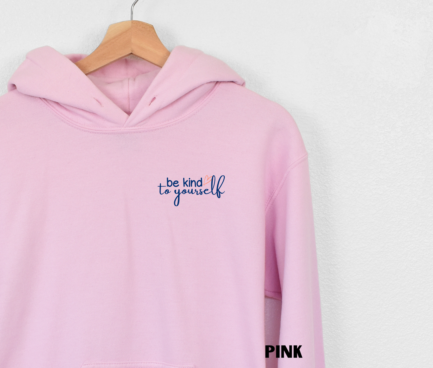 Close up of an embroidered Hoodie in light pink with "be kind to yourself' in navy blue text with a pink heart on the upper left chest. 