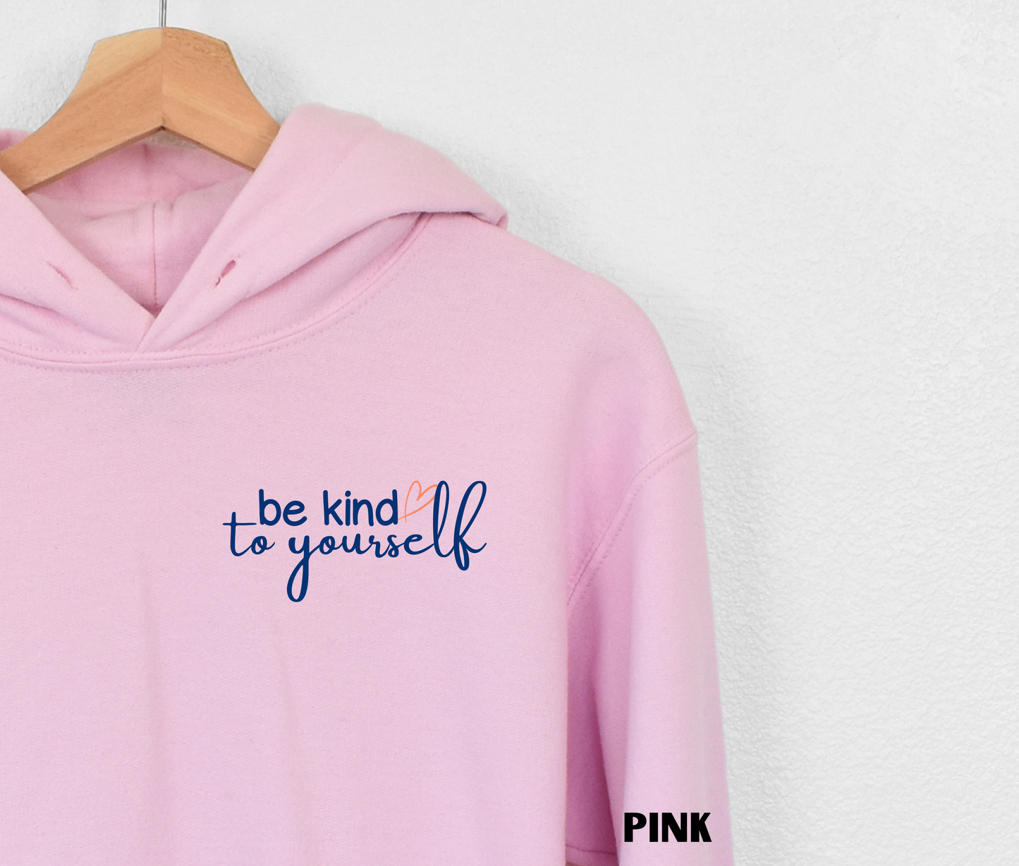 Close up of an embroidered Hoodie in light pink with "be kind to yourself' in navy blue text with a pink heart on the upper left chest. 