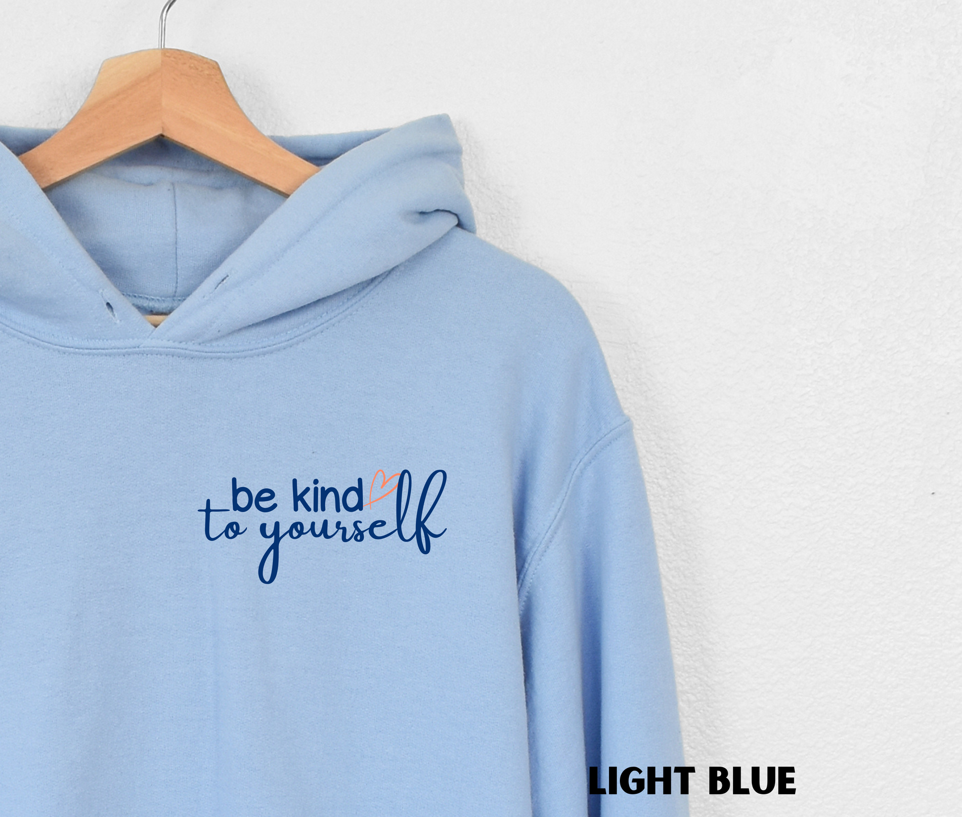 Close up of an embroidered Hoodie in light blue with "be kind to yourself' in navy blue text with a pink heart on the upper left chest. 