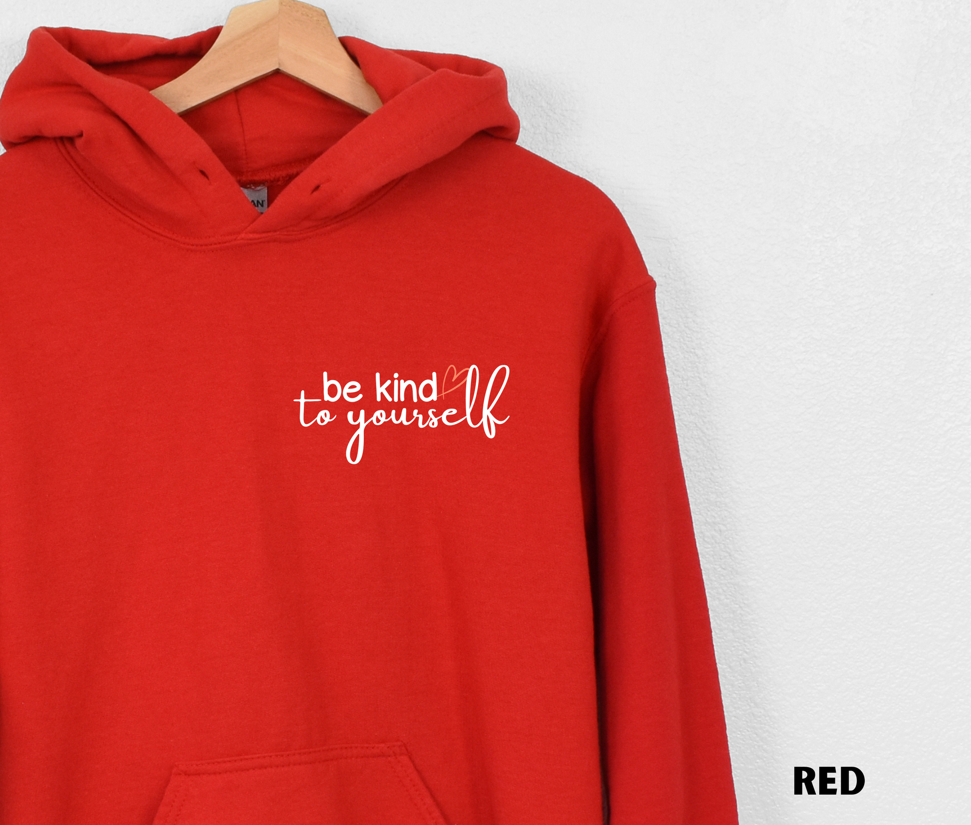 Close up of an Embroidered Hoodie in red with "be kind to yourself' in white text with a pink heart on the upper left chest. 