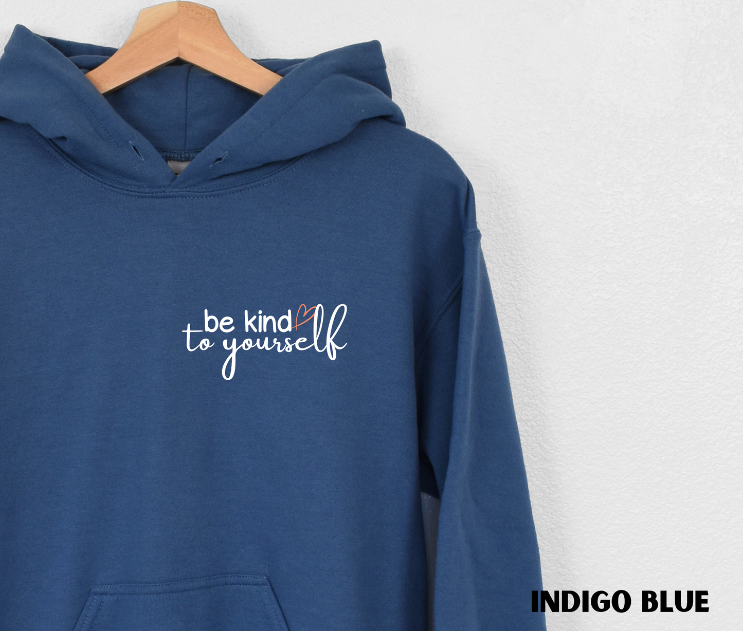 Close up of an Embroidered Hoodie in indigo blue with "be kind to yourself' in white text with a pink heart on the upper left chest. 