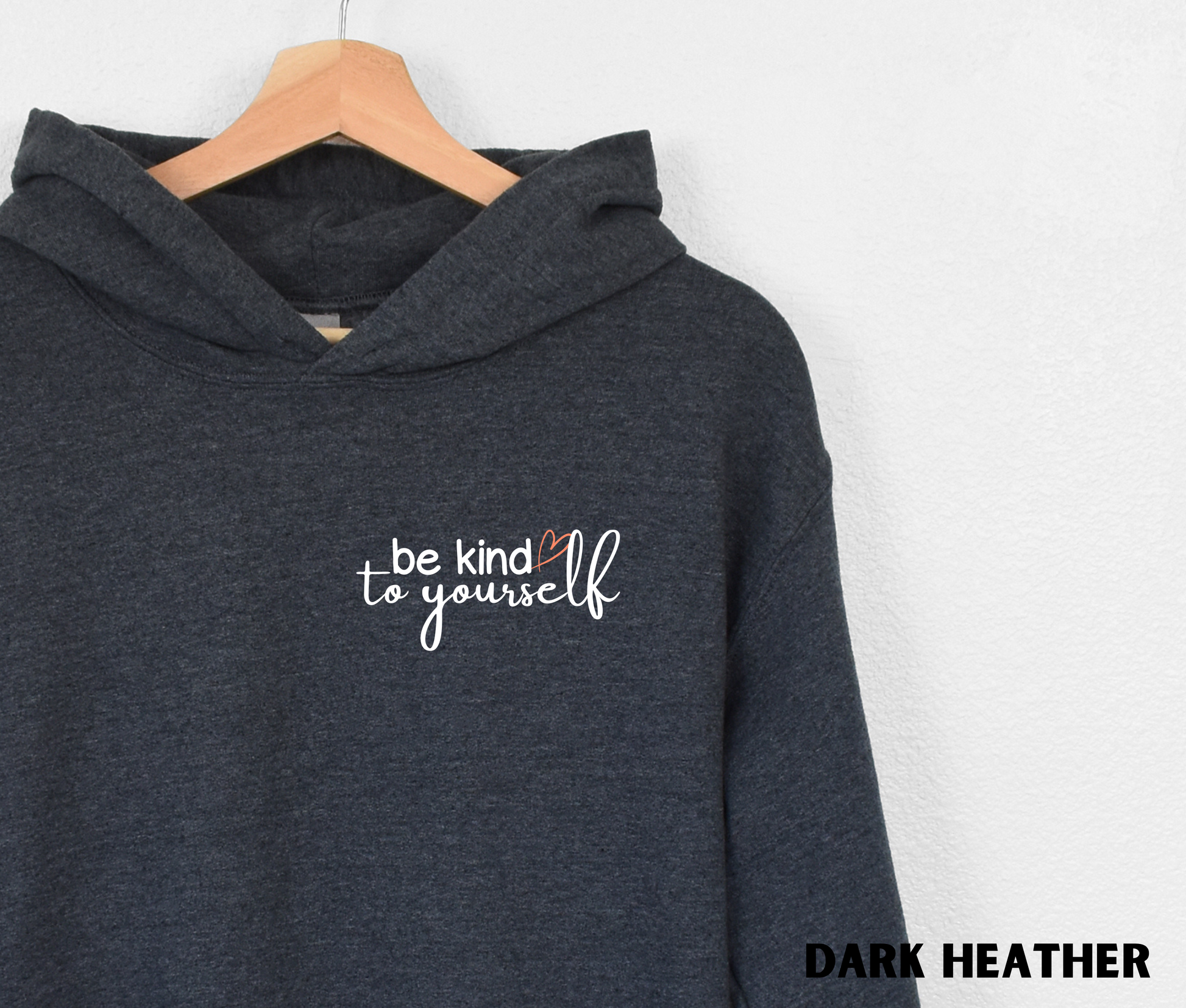 Close up of an Embroidered Hoodie in dark heather with "be kind to yourself' in white text with a pink heart on the upper left chest. 