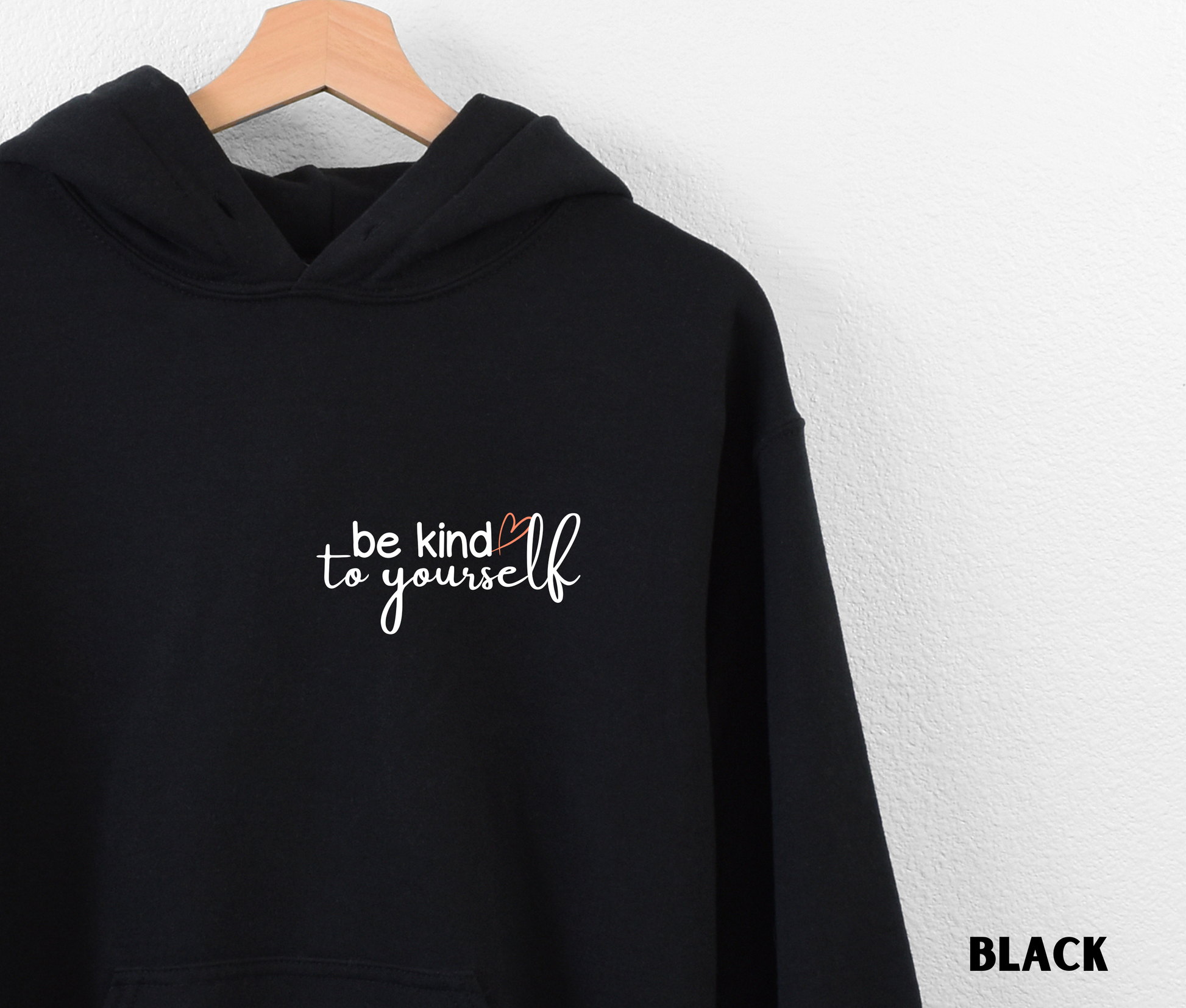 Close up of an Embroidered Hoodie in black with "be kind to yourself' in white text with a pink heart on the upper left chest. 