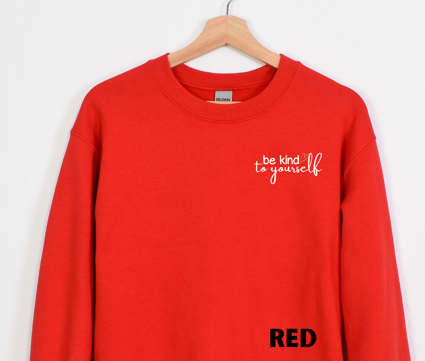 A red blue embroidered crewneck sweatshirt with the words "be kind to yourself" embroidered in white with a pink heart on the left chest.