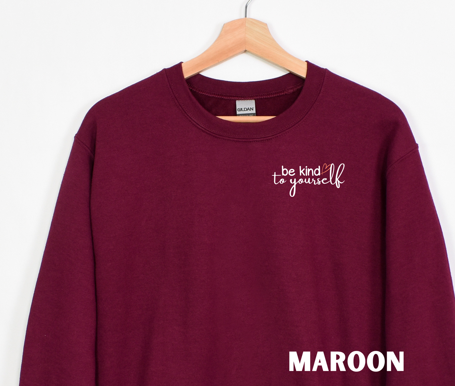 A maroon embroidered crewneck sweatshirt with the words "be kind to yourself" embroidered in white with a pink heart on the left chest.